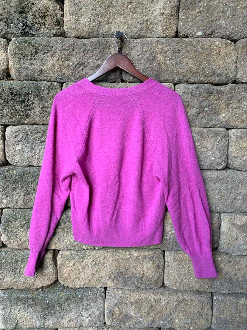 Bright pink long-sleeve sweater with ribbed cuffs and hem hanging on wooden hanger against stone wall.