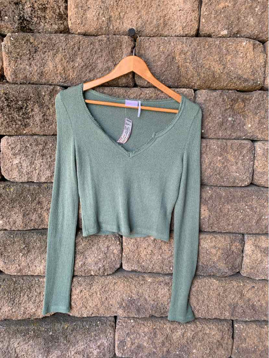 Sage green long-sleeve knit crop top with V-neck and slim fit hanging on wooden hanger against stone wall.