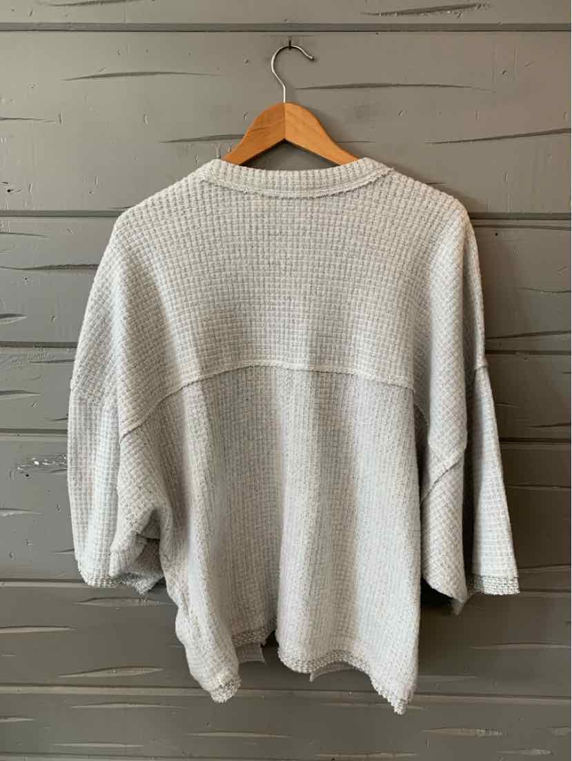 Light gray knitted sweater with dropped shoulders and loose three-quarter sleeves hanging on wooden hanger.