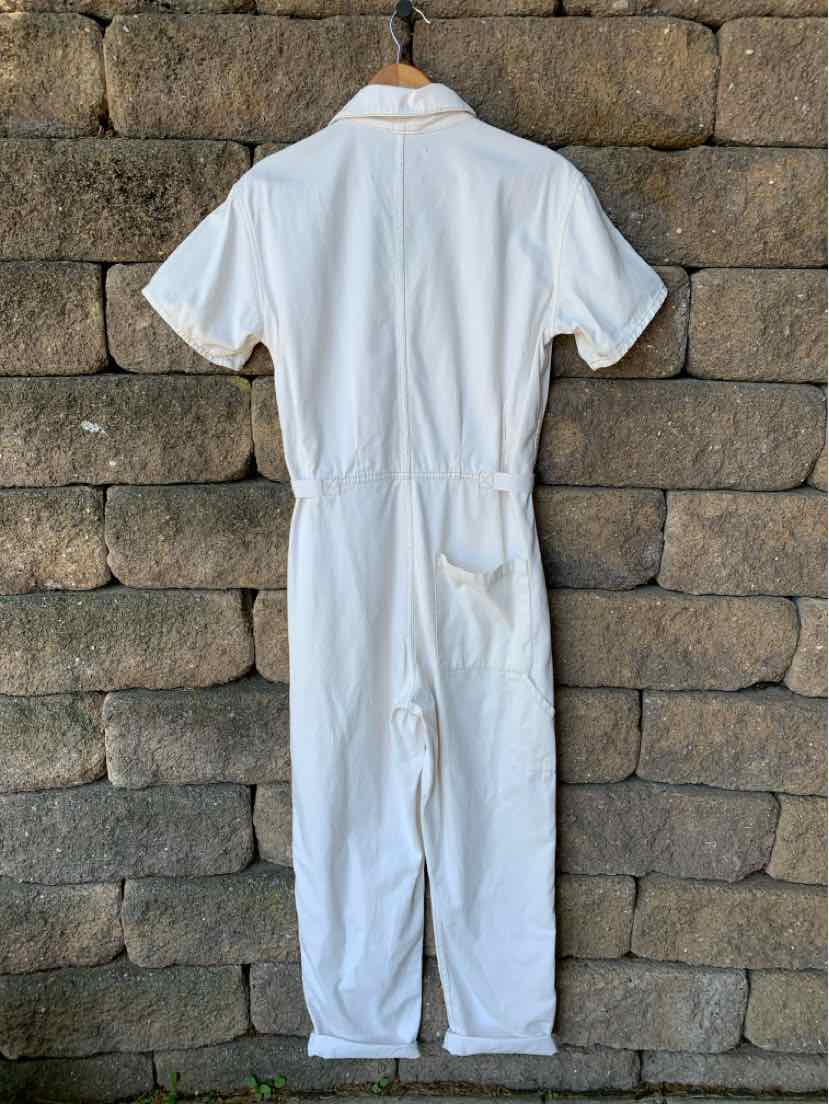 White short-sleeve coverall with cinched waist and back pocket on stone wall background.