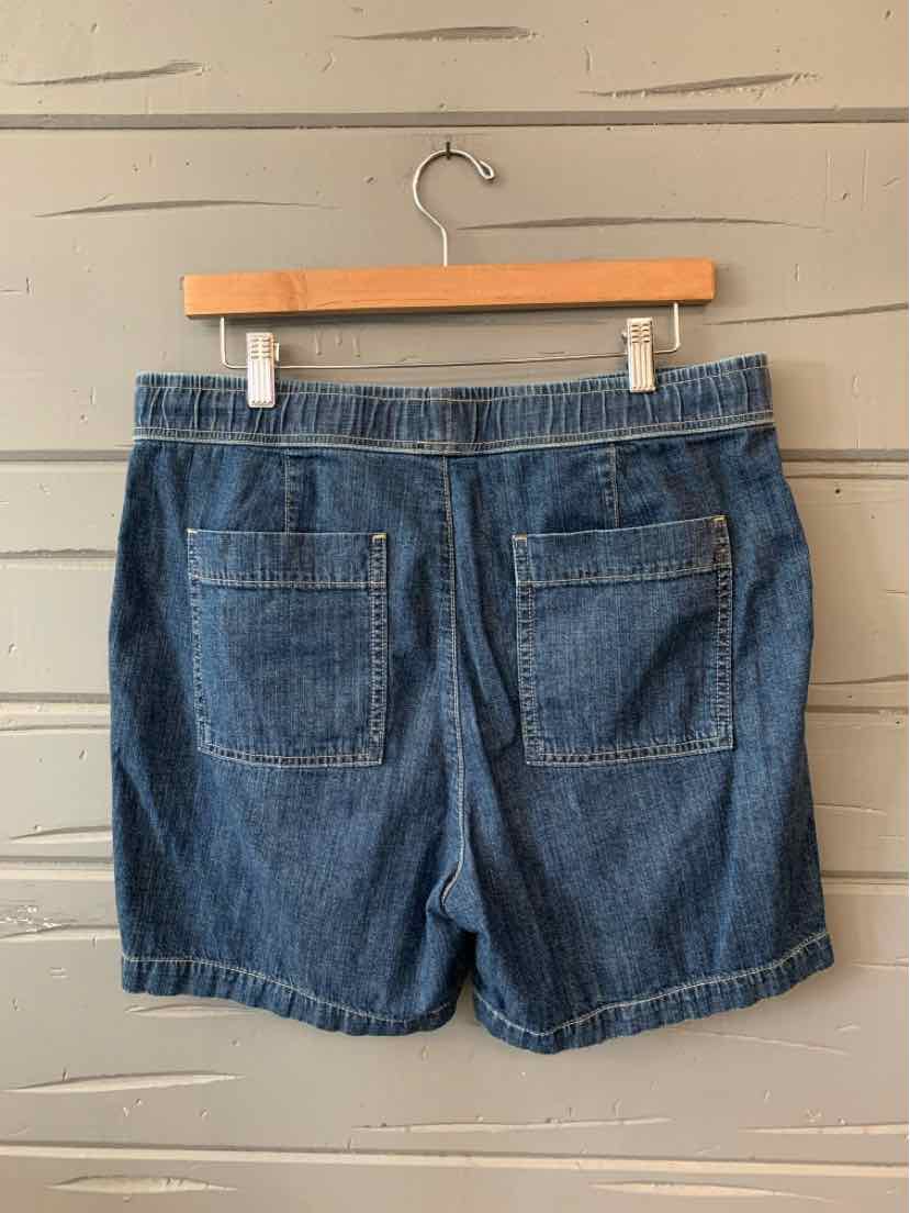 Dark blue denim shorts with two back patch pockets hanging on wooden hanger against gray wooden wall.