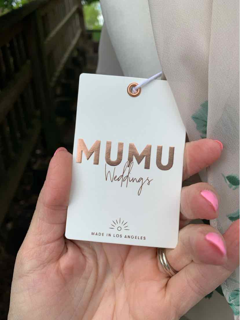 Hand holding beige Mumu Weddings clothing tag with rose gold foil lettering and pink nail polish.