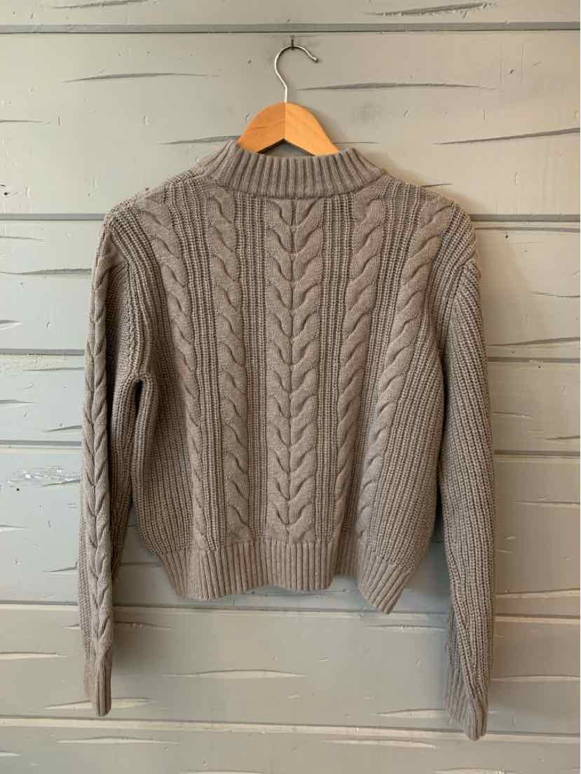Light gray cable knit sweater with long sleeves and ribbed hem on wooden hanger against gray wall.