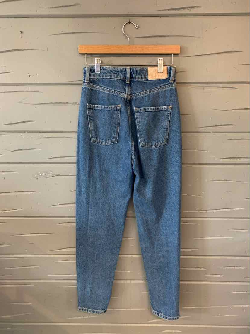 High-waisted medium blue denim jeans with back pockets and tapered legs hanging on wooden hanger against gray wall.