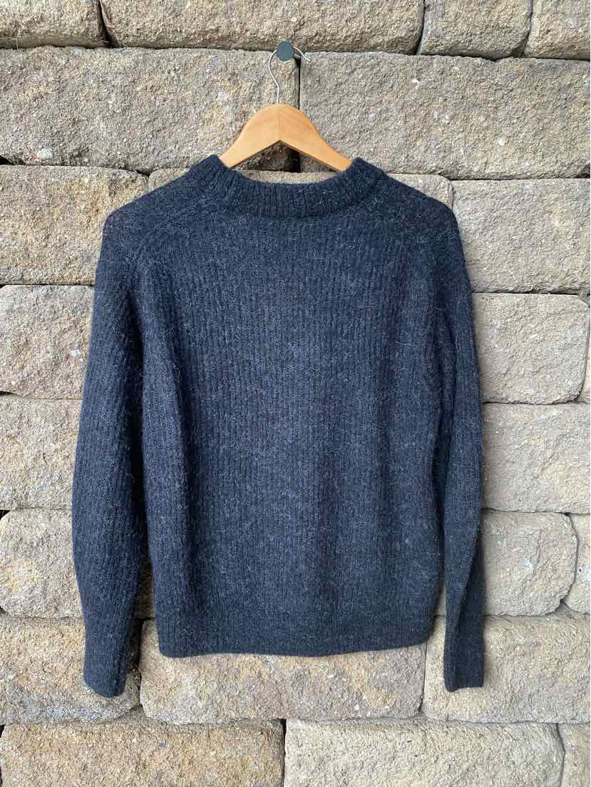 Dark gray knit sweater with long sleeves and a crew neckline hanging on a wooden hanger against a stone wall background.