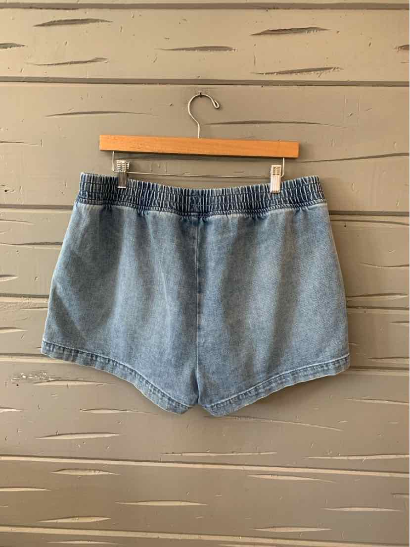Light blue denim shorts with elastic waistband and a relaxed fit hanging on a wooden hanger against a beige wall.