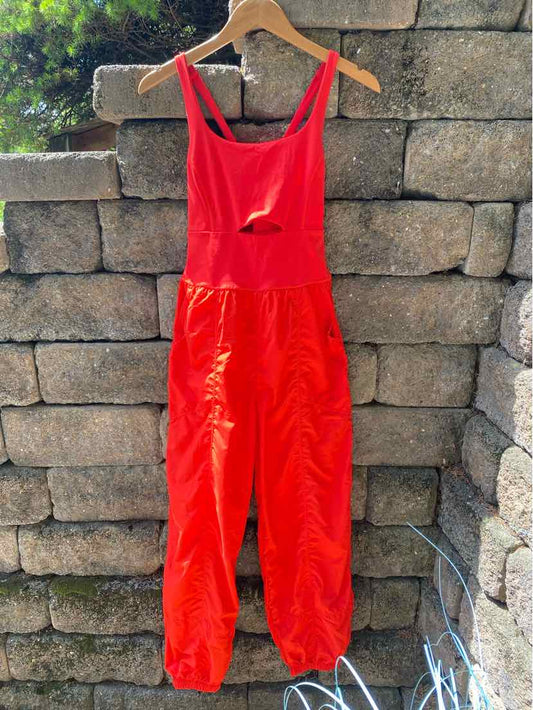 Red sleeveless jumpsuit with adjustable straps and gathered ankle cuffs hanging outdoors on wooden hanger.