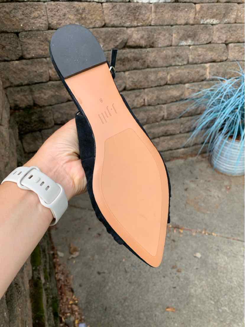 Black pointed-toe flat shoe with tan sole and a slight heel, held outdoors against stone wall background.