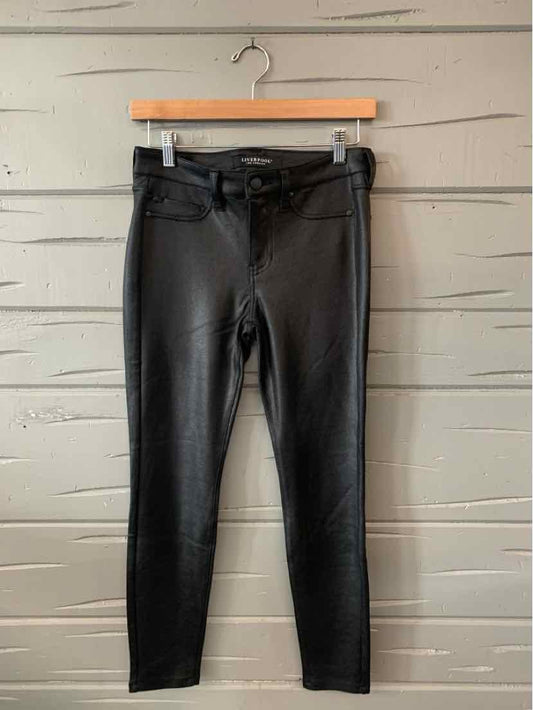 Black slim-fit pants with front pockets and button closure hanging on wooden hanger against gray wooden wall.