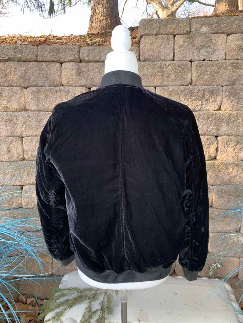 Black velvet bomber jacket with ribbed cuffs and waistband on white mannequin against stone wall background.