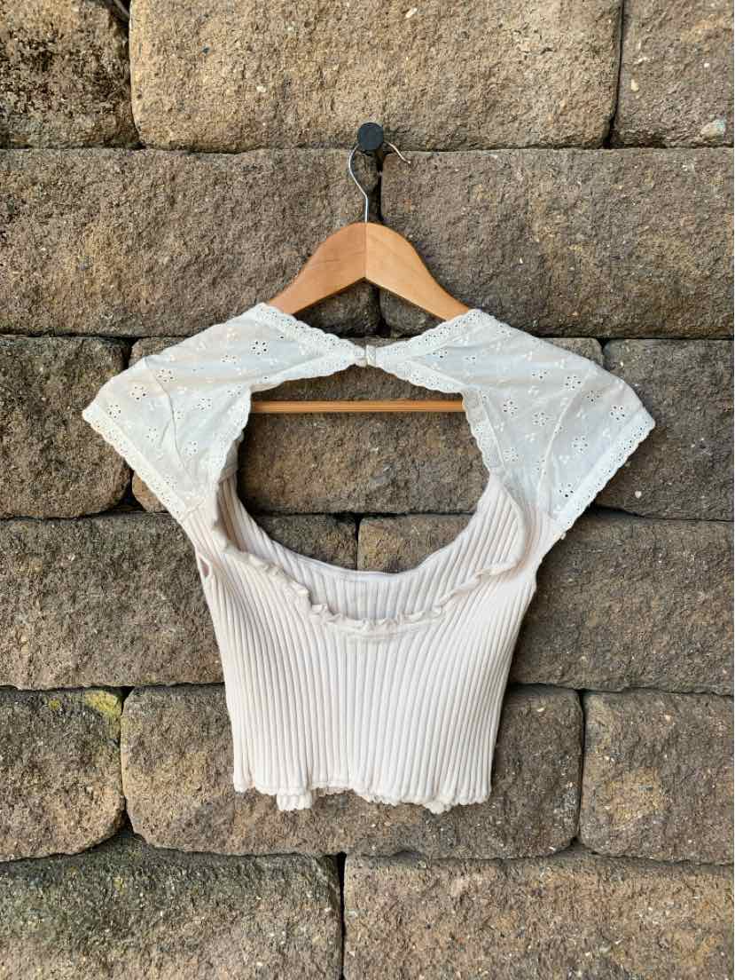 Tan ribbed crop top with scalloped edges and fitted short sleeves on wooden hanger against stone wall.