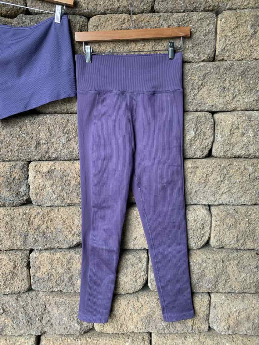 Purple high-waisted seamless leggings with a wide waistband hanging on a wooden hanger against a stone wall.