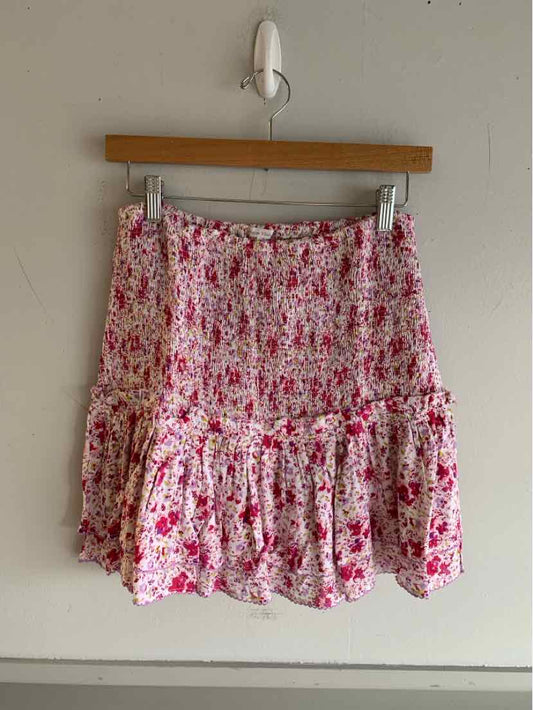 Pink floral mini skirt with ruffled hem and smocked waistband hanging on wooden hanger against beige wall.