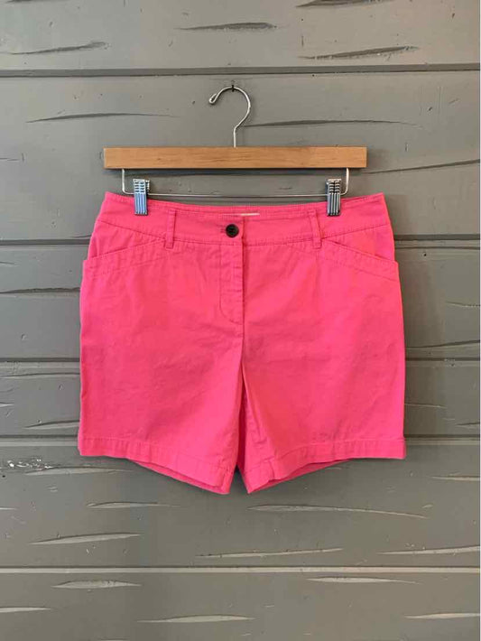 Bright pink tailored shorts with front button and zipper, side pockets, and belt loops on wooden hanger against gray wall.