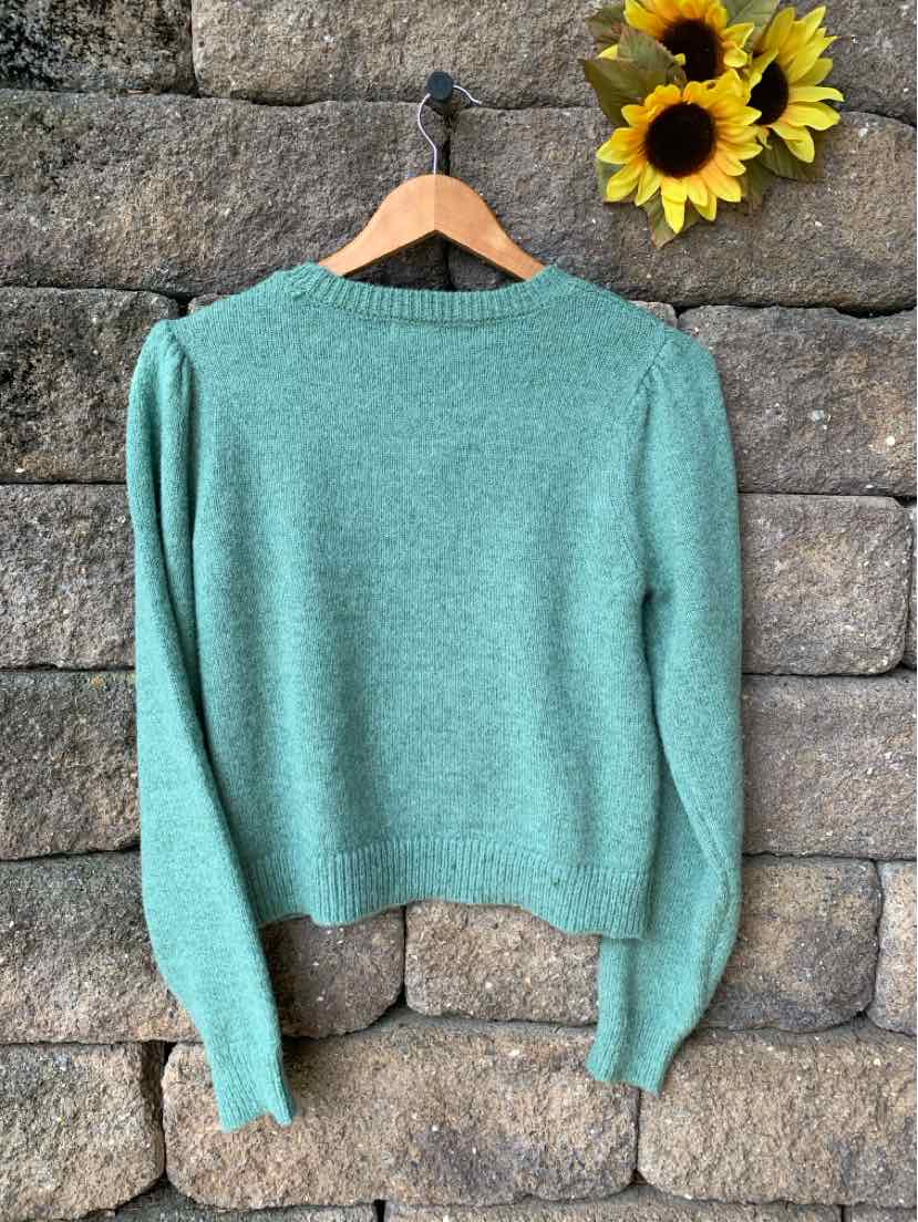 Light teal-green knit sweater with long sleeves and a rounded neckline, displayed on a wooden hanger against a stone wall.
