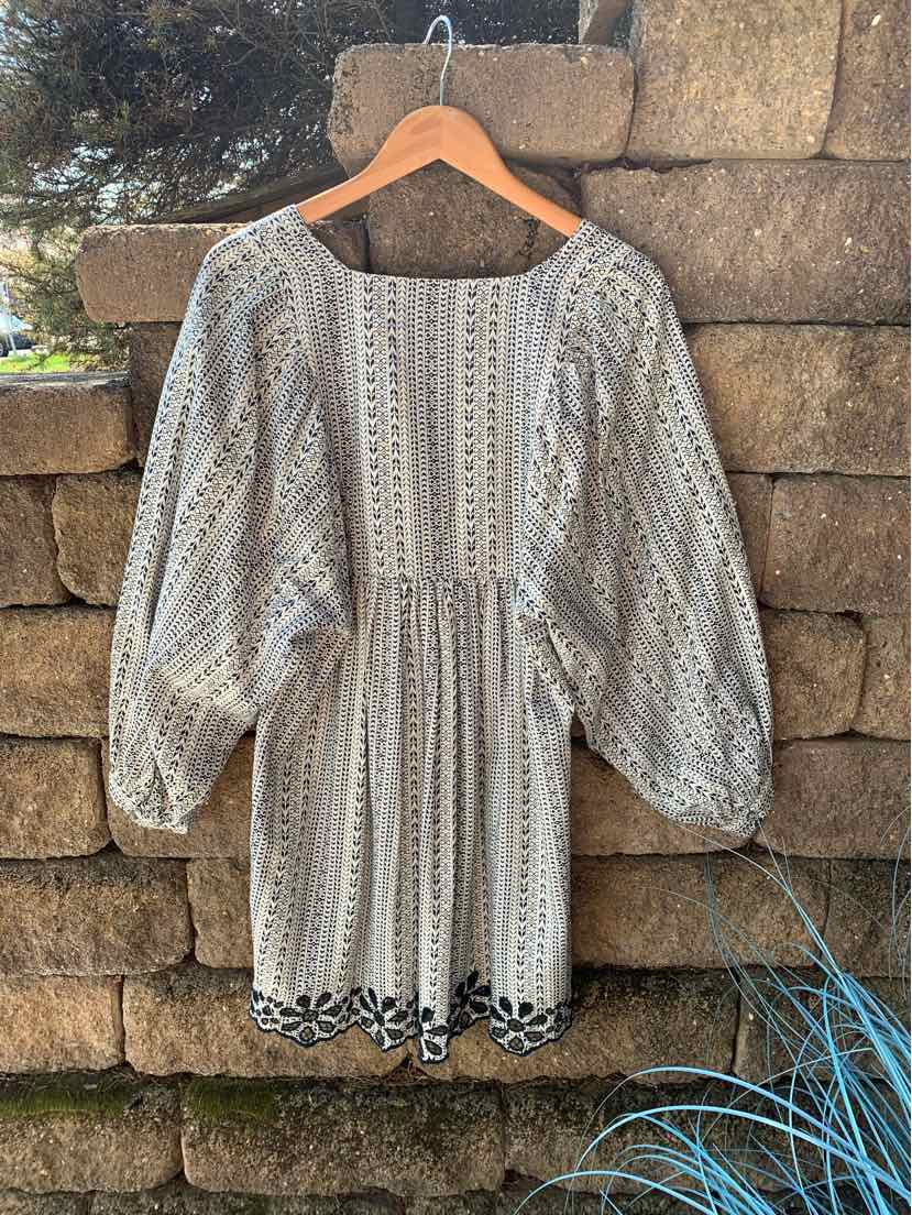 Cream and black patterned dress with puffed long sleeves and cinched waist on wooden hanger against stone wall.