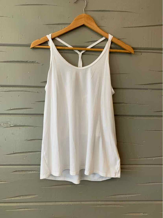 White sleeveless tank top with thin straps and racerback detail hanging on wooden hanger against gray wooden wall.