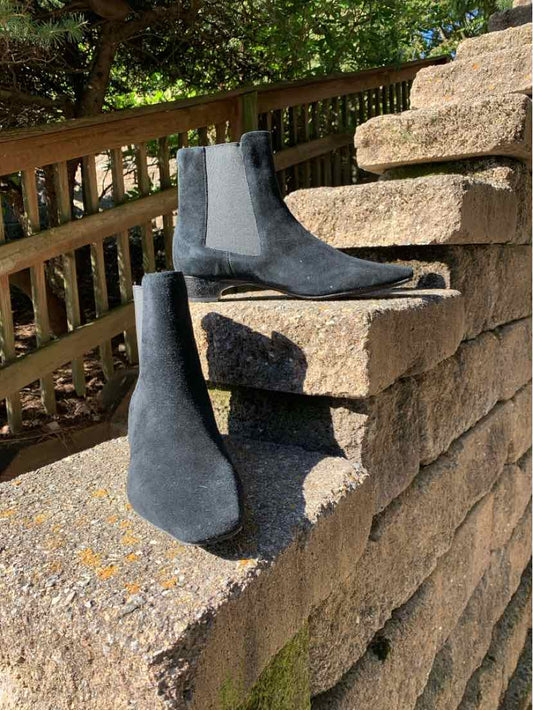 Black suede ankle boots with pointed toes, low heels, and sleek pull-on shafts displayed on a stone wall.