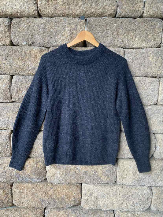 Dark gray knit sweater with round neckline and long sleeves hanging on wooden hanger against stone wall background.