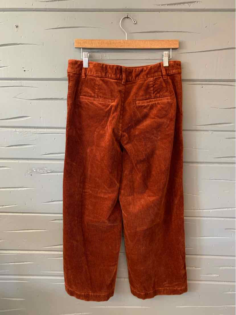 Rust-colored corduroy pants with a relaxed, straight-leg fit hanging on a wooden hanger.