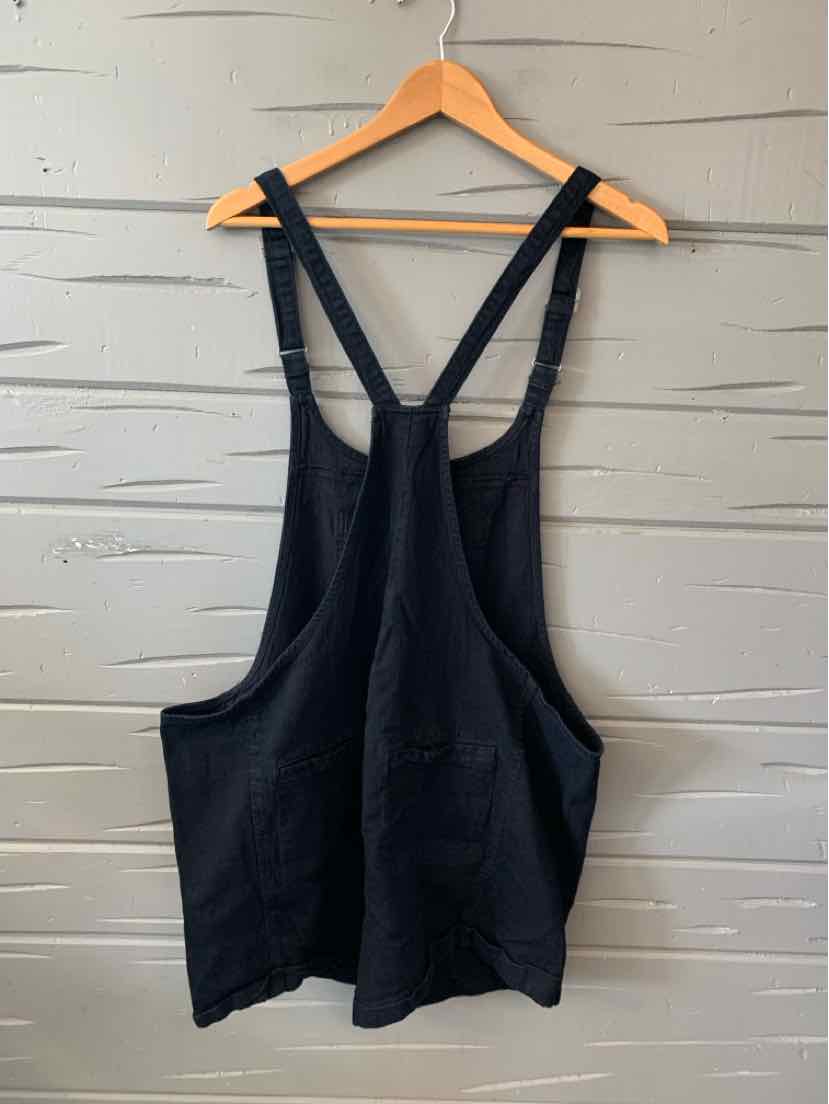 Black sleeveless apron with wide shoulder straps and front pockets hanging on wooden hanger.