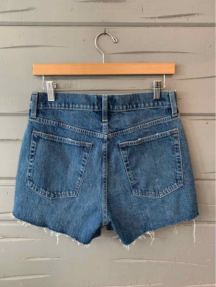 Medium blue denim shorts with a frayed hem and two back patch pockets hanging on a wooden hanger.