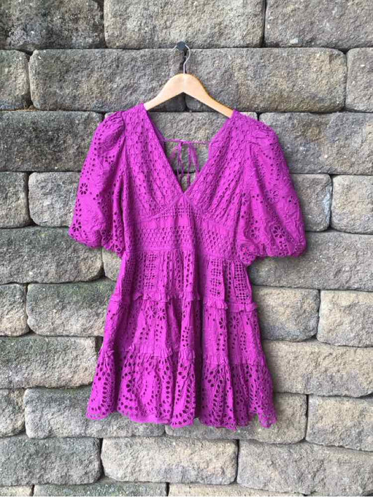 Purple eyelet lace dress with puff sleeves and tiered skirt on wooden hanger against stone wall.