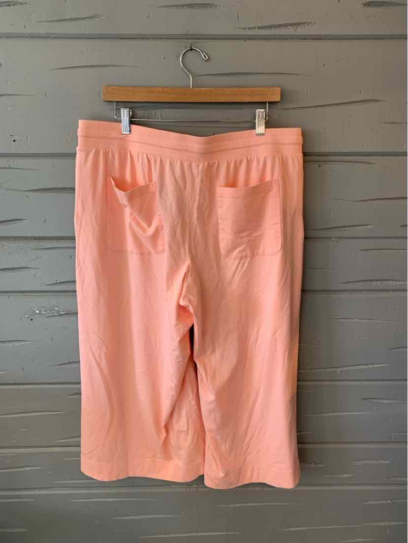 Coral peach knee-length shorts with elastic waistband and two front pockets hanging on wooden hanger against gray wooden wall.