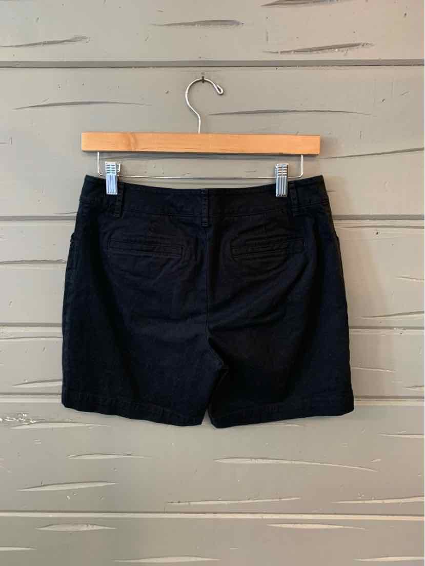 Black shorts in size 6P with back pockets and belt loops hanging on a wooden hanger against a gray wooden wall.