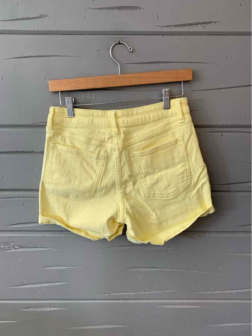 Light yellow women's shorts with an elastic waistband and two back pockets hanging on a wooden hanger against a gray wall.