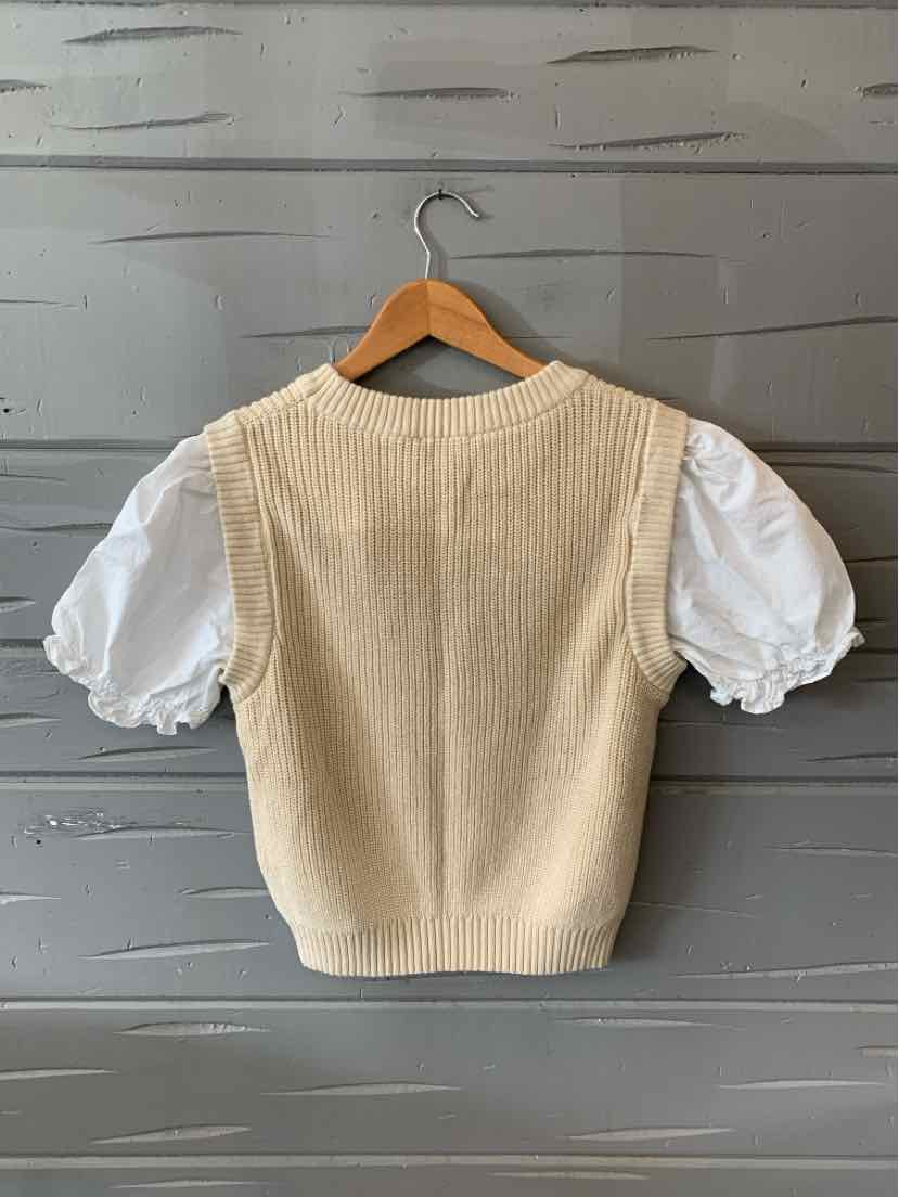 Beige knit vest layered over a white puff-sleeve blouse on a wooden hanger against a gray wooden wall.