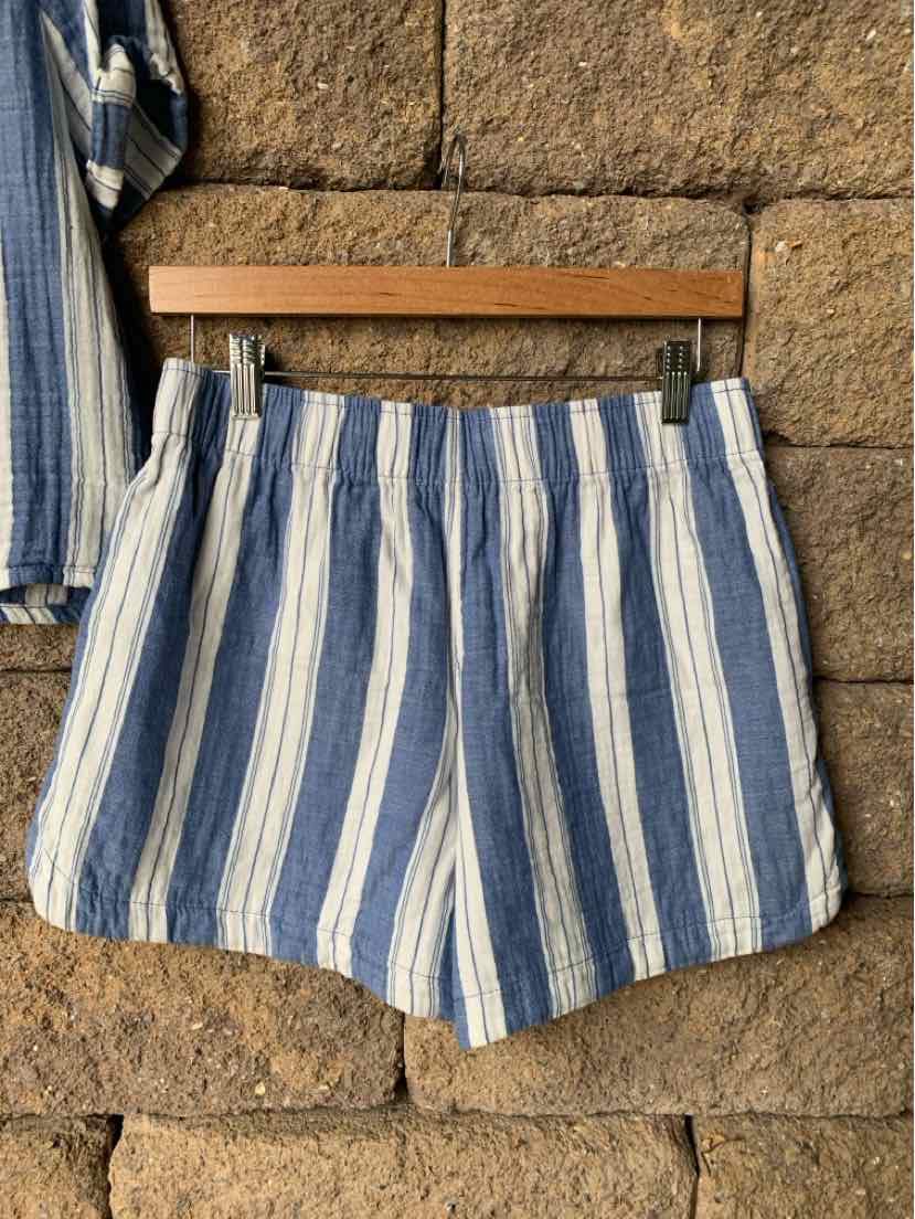 Blue and white vertical striped casual shorts with elastic waistband hanging on wooden hanger against stone wall.