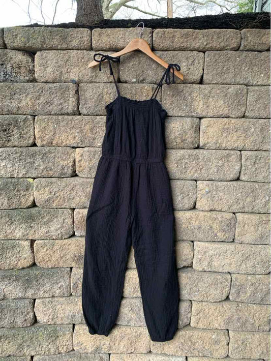 Black sleeveless jumpsuit with adjustable tie straps and elastic cuffs hanging on wooden hanger against stone wall.