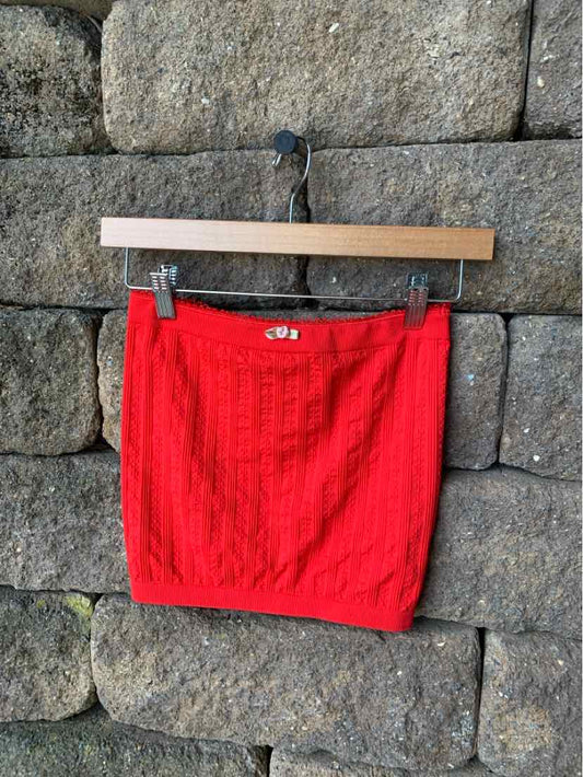 Red knit mini skirt with textured vertical cable pattern, hanging on wooden hanger against stone wall background.