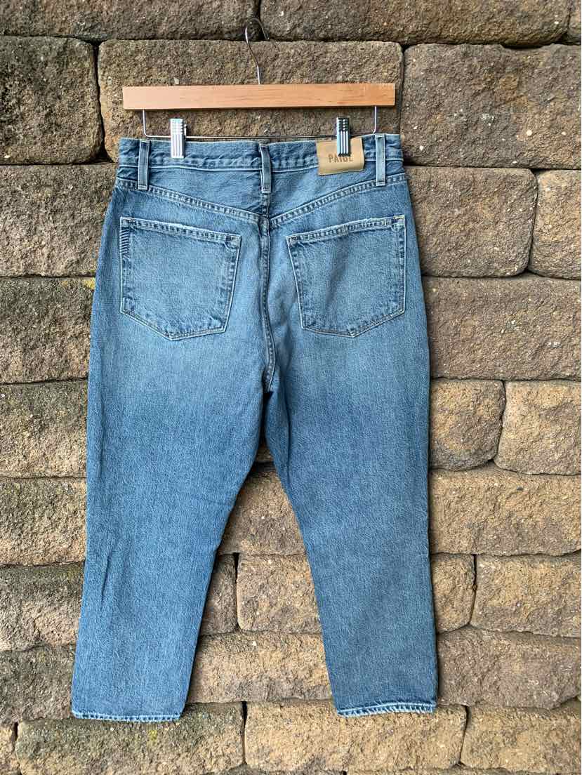 High-waist light blue denim jeans with straight legs and two back pockets hanging on wooden hanger against stone wall.