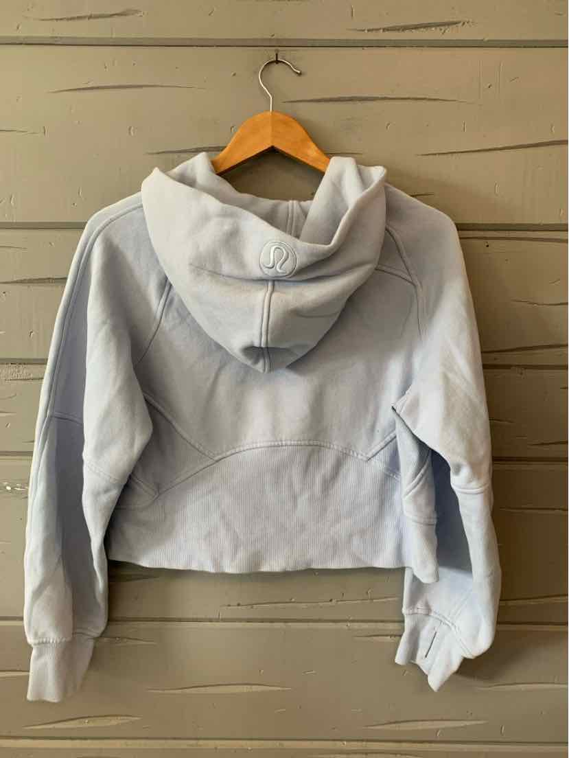 Light gray cropped hoodie with long sleeves and a front pocket hanging on a wooden hanger against a beige wooden wall.