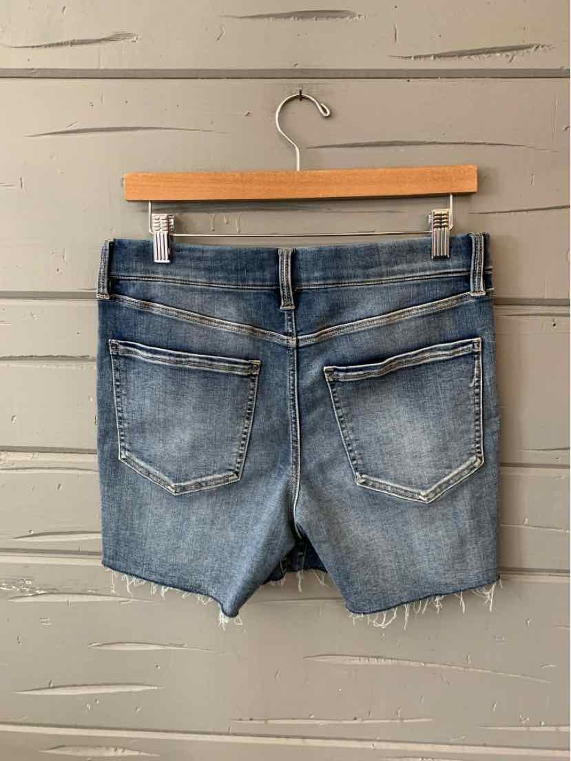 Blue denim shorts with frayed hems and two back pockets hanging on wooden hanger against gray wood background.