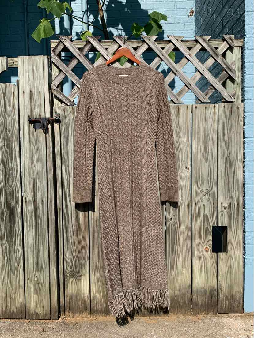 Beige knitted long-sleeve dress with cable pattern and fringe hem hanging on wooden gate.