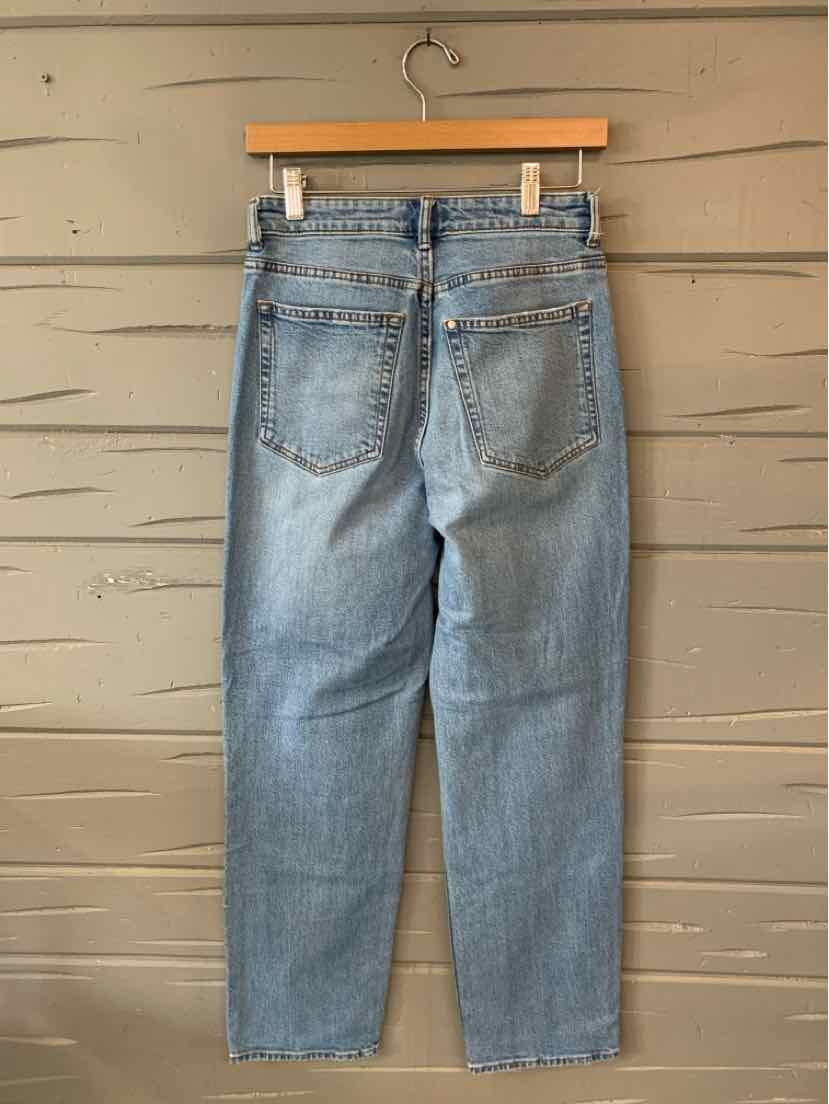 Light blue denim jeans with a straight leg cut and two back pockets hanging on a wooden hanger against a beige wooden wall.