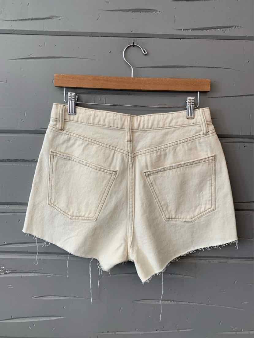 Off-white denim shorts with frayed hems and two back pockets on wooden hanger against gray wall.
