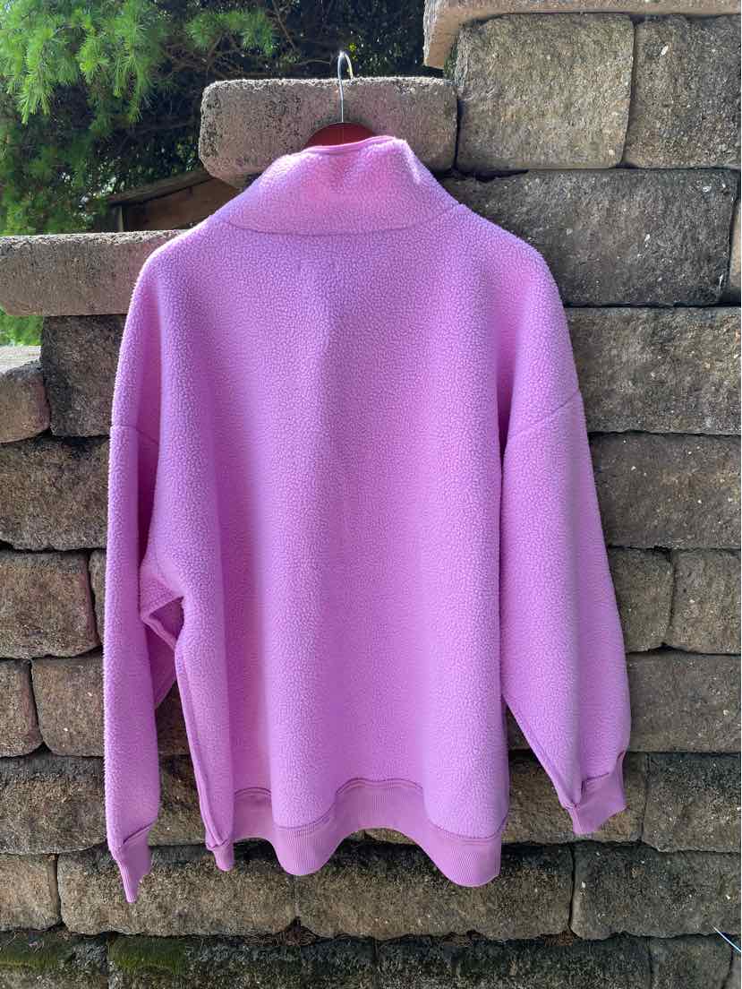Lavender oversized sweatshirt with high collar and dropped shoulders hanging against stone wall background.