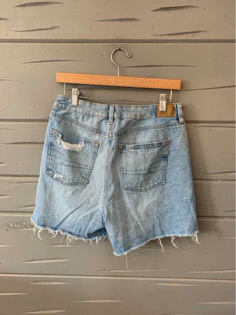 Light blue denim shorts with frayed hems and back pockets on a wooden hanger against a gray wooden wall.