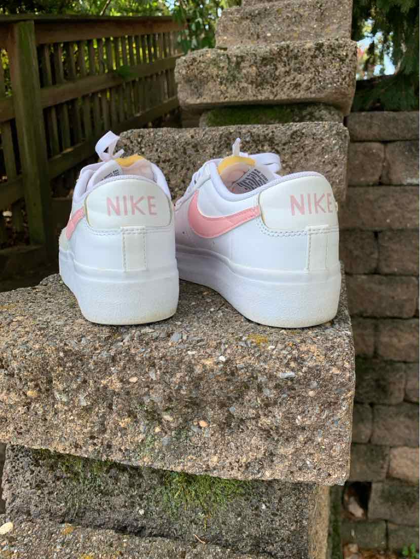 White Nike sneakers with pale pink Swooshes and heel lettering, shown from the back with chunky rubber soles.