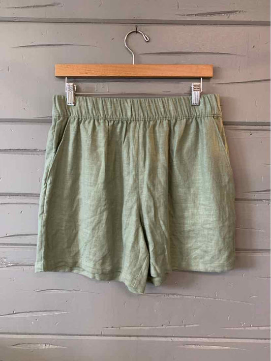 Light olive green casual shorts with elastic waistband and side pockets hanging on wooden hanger against gray wood.