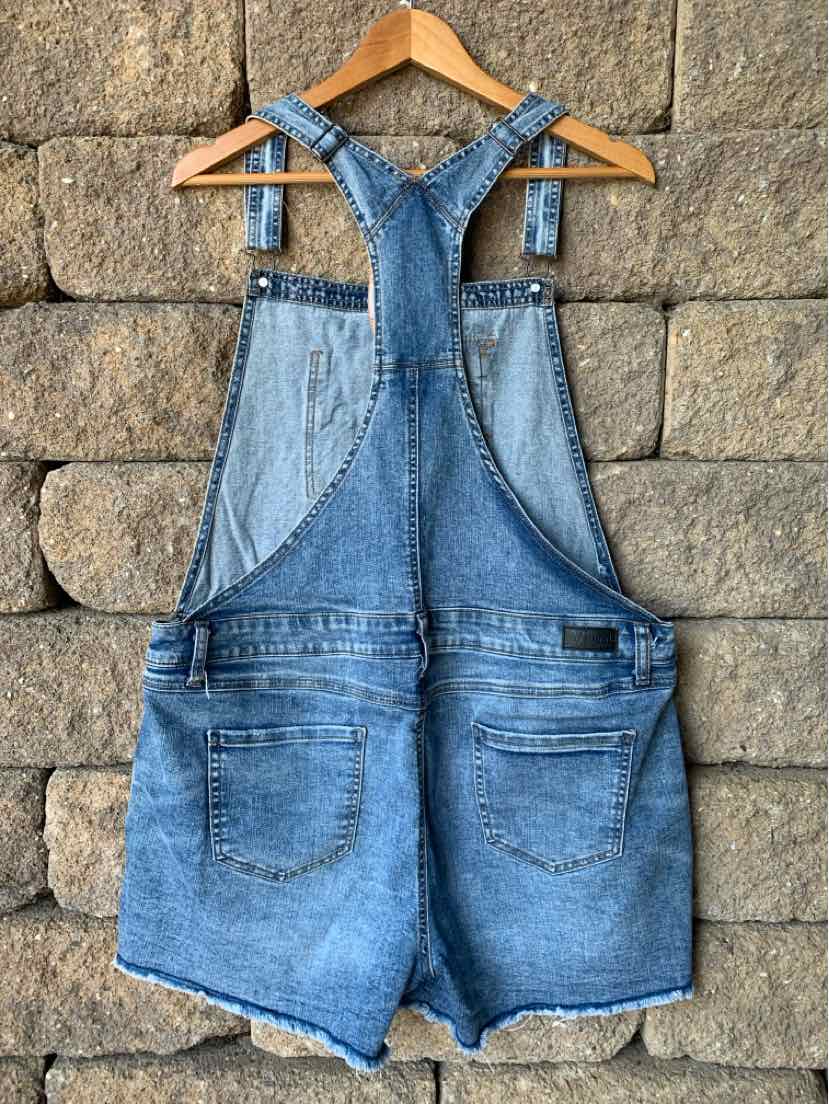 Blue denim overall shorts with adjustable shoulder straps and back pockets hanging on wooden hanger against stone wall.