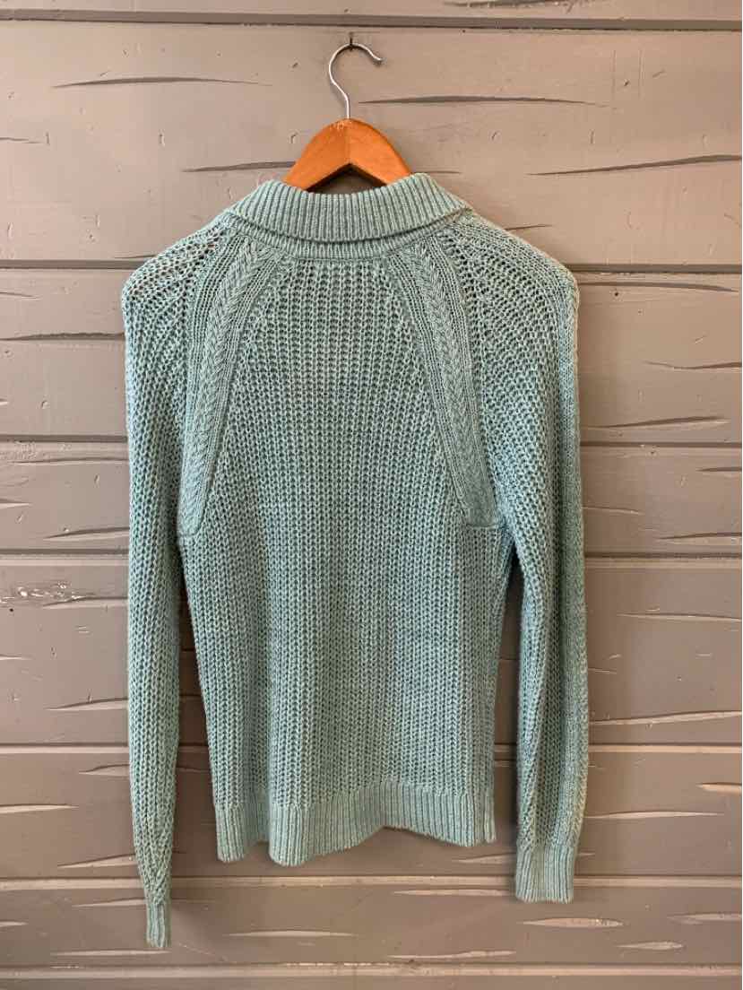 Light blue knit sweater with ribbed collar and long sleeves hanging on wooden hanger against wooden wall.