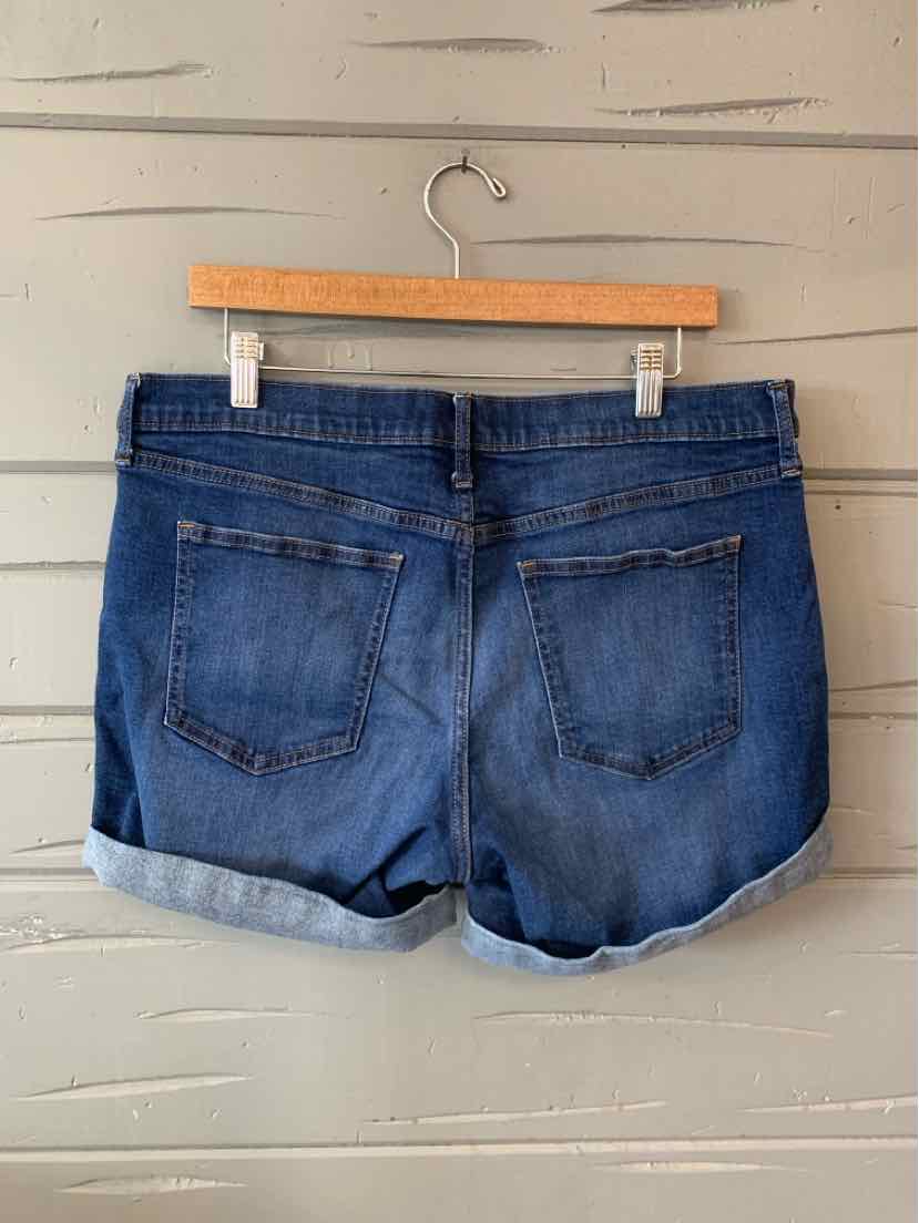 Dark blue denim shorts with rolled cuffs and two back pockets hanging on a wooden hanger against a gray wooden wall.