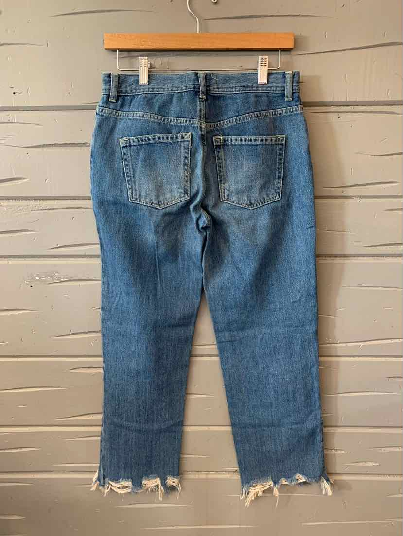 Blue denim cropped jeans with frayed hems and back patch pockets hanging on wooden hanger against beige wall.