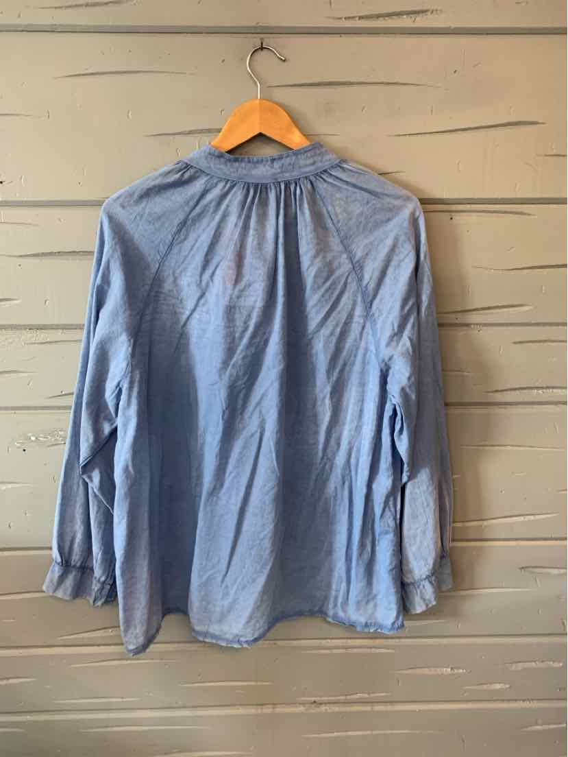 Light blue long-sleeve shirt with gathered yoke and buttoned cuffs hanging on wooden hanger against wooden wall.