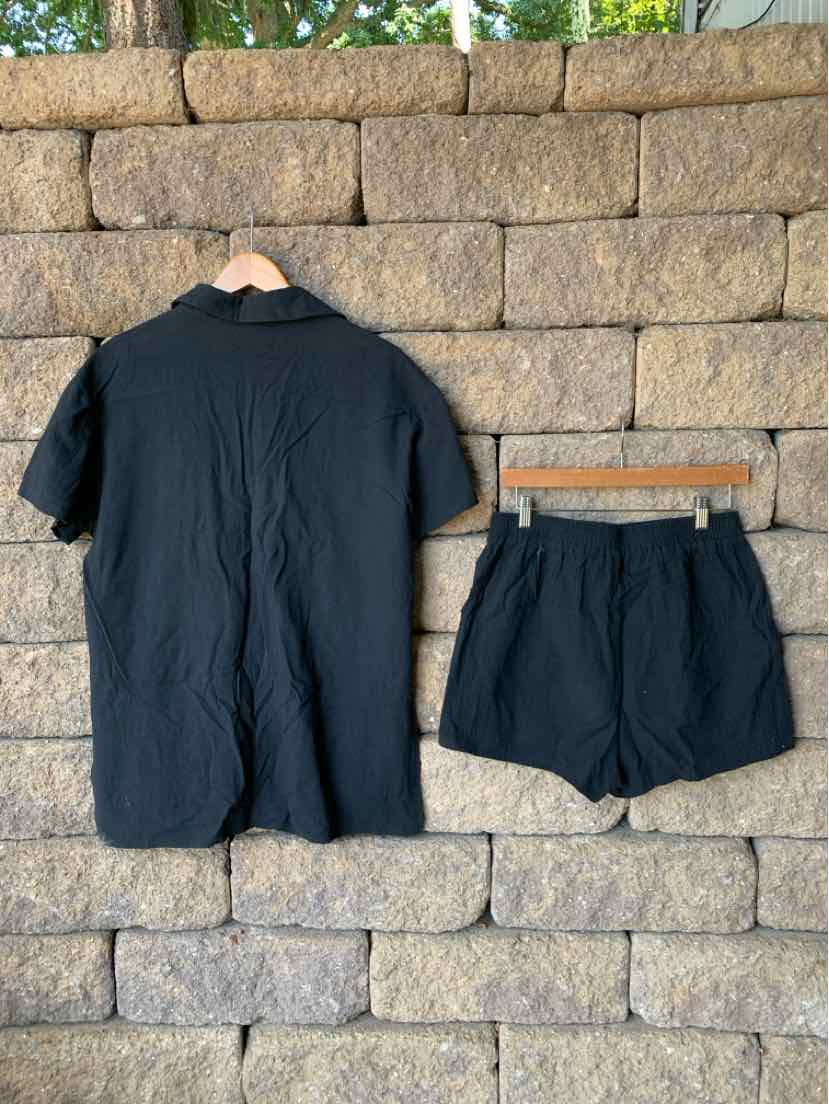 Black short-sleeve button-up shirt and matching black shorts set hanging on wooden hangers against a stone wall background.